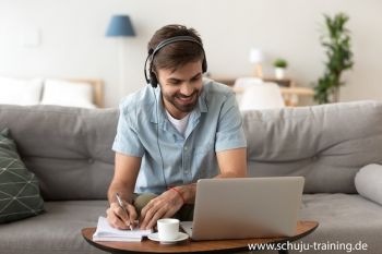Ein Mann sitzt auf einem Sofa, trägt Kopfhörer und sieht auf einem Laptop auf dem Tisch vor ihm